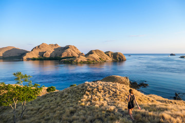 Naklejka premium A woman standing on top of a small island, enjoying the morning sun over Komodo National Park, Flores, Indonesia. Golden hour over the islands and sea. Some boats anchored to the bay. New day 
