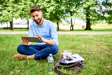 Handsome young man sitting on grass in park with tablet during lunch break