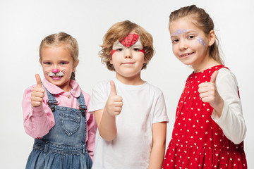 cheerful friends with colorful face paintings showing thumbs up while looking at camera isolated on white © LIGHTFIELD STUDIOS