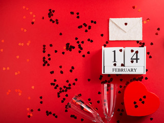Happy valentines day concept flat lay with champagne flutes and red heart confetti on red background, wooden calendar, envelope, heart shaped gift box