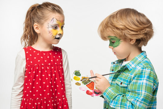 Adorable Boy With Painted Gecko Mask Holding Palette And Paintbrush Near Friend With Tiger Muzzle Painting On Face Isolated On White