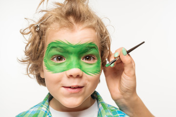 cropped view of artist painting gecko mask on face of shaggy, cheerful boy isolated on white