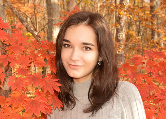 Young Russian pretty girl in gray sweater smiling and looking at the camera on autumn background with red maple leaves on a sunny warm day of October