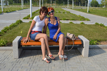 Two forty year old sisters sincerely smiling with teeth and sitting on the bench on a sunny summer...