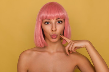 Obraz premium Close-up of young pretty blue-eyed pink haired woman with trendy bob haircut pouting her lips while looking at camera and keeping index finger on her cheek, isolated over mustard background