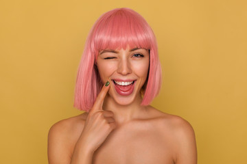 Joyful young attractive pink haired female with bob hairstyle holding index finger on her face...
