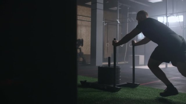 Young strong athletic Caucasian man exercising with heavy weight training sled towards camera in large gym slow motion