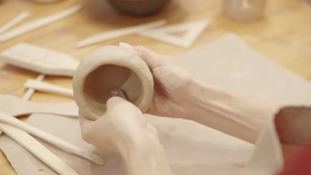 Over-shoulder close-up shot of hands of unrecognizable female potter sitting in workshop, making clay bowl, pinching and forming its walls, and smoothing surface, and selection of tools lying on table