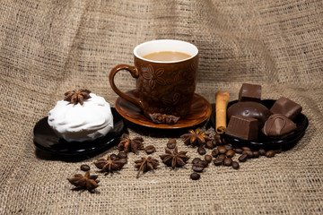 On the table is a cup of coffee with milk, a saucer with chocolates and marshmallows. Nearby are aniseles, coffee beans and cinnamon sticks.