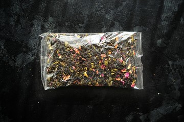 Black tea in a plastic transparent bag. Tea with flowers and packaging on a black background isolated. View from above.
