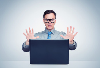 Frightened man in a shirt, tie and glasses raised his hands in front of a laptop. Fear emotion...