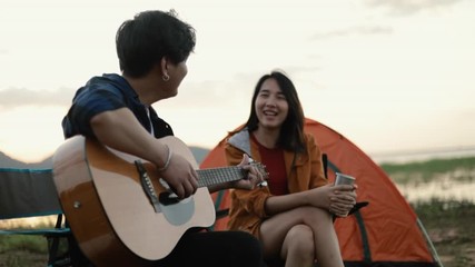 A happy young asian couple backpacker playing the guitar and singing a song together beside camping relaxing on vacation. - Powered by Adobe