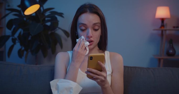 Close up of young woman wiping tears from her face with paper tissue while looking at phone screen. Upset girl cant believe bad news reading on her smartphone and crying.
