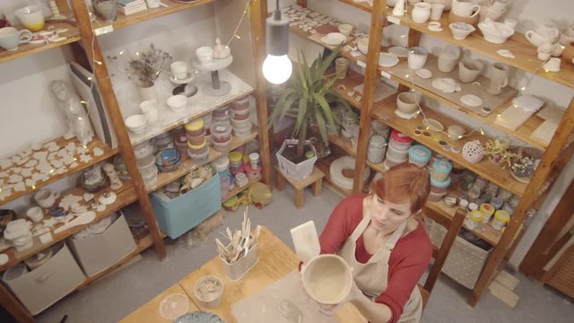 Top shot of Caucasian female ceramics artist making earthenware bowl in studio, patting and smoothing outer surface with wooden paddle, and shelves with semifinished produce and supplies in background