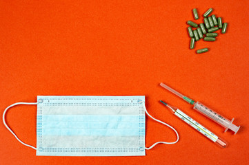 Sterile medical mask, mercury thermometer, syringe, capsules with medicine on an orange background. Virus and disease protection