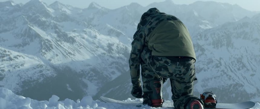 Back view of a professional snowboarder buckling into bindings on a mountain peak early in the morning, preparing for freeride snowboarding. 4K UHD, shot with anamorphic lens