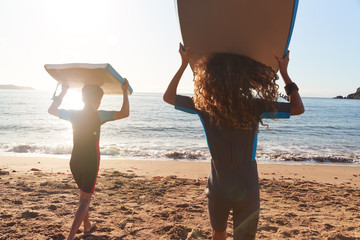 Rear View Of Children In Wetsuits Carrying Bodyboards On Summer Beach Vacation Having Fun By Sea