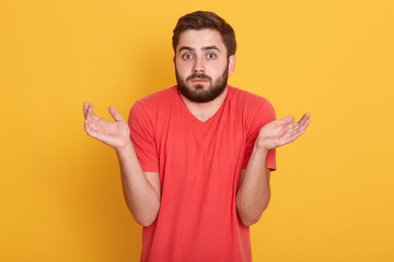 Fototapeta premium Doubtful emotional handsome young man standing isolated over yellow background, raising hands, making gesture, having confused facial expression, wearing red casual t shirt. Emotions concept.