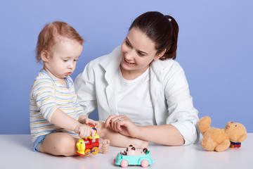 Obraz na płótnie Canvas Horizontal shot of mother and child son being engaged in creativity in studio over blue background, mummy looking smiling at her charming child, cute kid playing with car toys. Motherhood concept.