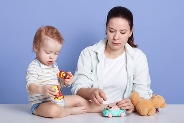 Horizontal shot of mom with kid son play together in room at home, infant wearing striped bodysuit, dark haired mother dresses casual clothing, family posing isolated over blue studio background.