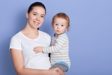 Studio shot of beautiful young mother standing and holding her baby boy in her arms in front of blue background, lady with dark hair and ponytail with cute infant, looking ar camera and smiling.