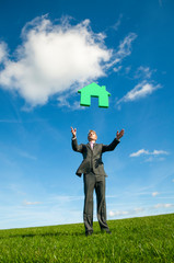 Businessman running outdoors on a meadow chasing a green house floating in blue sky