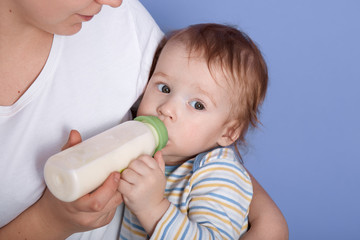 Hirizontal shot of liitle charming baby drinking milk of bottle, being in mother's arms, feels hunger, looking aside, wearing stripped shirt, infant relaxing and feeling happyness near her mommy.