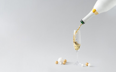 Champagne bottle pouring golden glitter into the glass with white and golden decoration against grey background. Celebration minimal Christmas party.