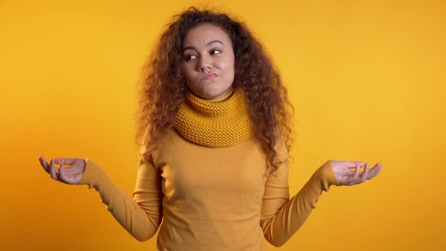  Young curly unsure girl shrugs her arms, makes gesture of I don't know, care, can't help anything .Young curly woman with trendy total warm look