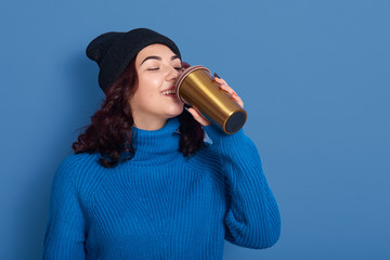Image of delighted cute satisfied young female drinking coffee from thermo mug, taking sip of hot...