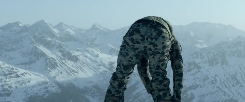 Back view of a professional snowboarder buckling into bindings on a mountain peak early in the morning, preparing for freeride snowboarding. 4K UHD, shot with anamorphic lens
