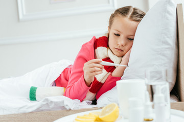sick upset child with fever in scarf looking on thermometer while lying on bed with medicines near