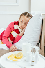 Obraz na płótnie Canvas sad ill kid in scarf lying on bed with medicines near