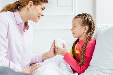 smiling mother giving hot drink to sick daughter in bed