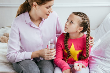 smiling mother giving glass of water to diseased daughter with pill