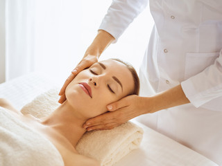 Obraz na płótnie Canvas Beautiful woman enjoying facial massage with closed eyes in spa center. Relaxing treatment concept in medicine