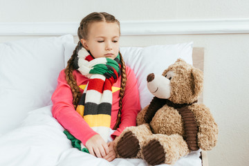 sad diseased kid in scarf sitting on bed with teddy bear