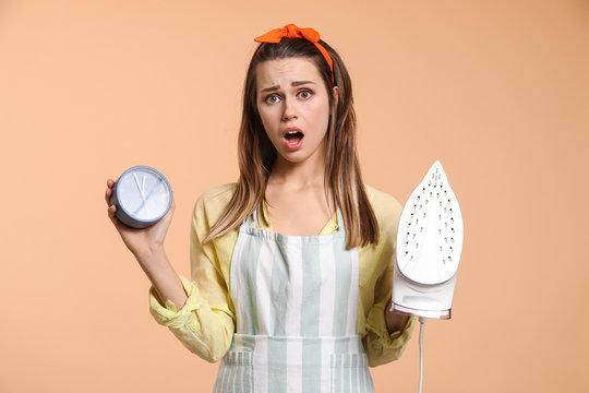 Stressed young housewife with iron and alarm clock on color background