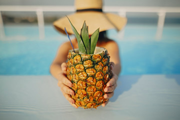 Delicious cocktail in pineapple. Summer vacation. Beautiful young woman in hat relaxing in pool with drink. Girl enjoying warm sunshine in pool on rooftop in luxury tropical resort.