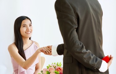 A man in black suit hiding a red box of wedding ring behind his back to propose to his girlfriend in pink dress