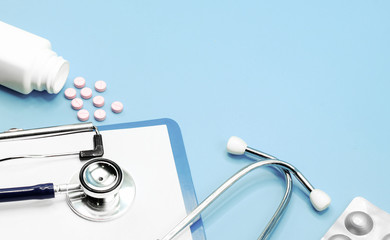 Flat lay of pills, medicines, stethoscope and writing pad paper clip board, on light blue background with copy space, healthcare and medical concept, top view photo.