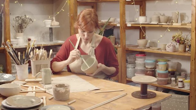 Dollying shot of Caucasian female potter with red hair working in ceramics studio, sitting at table and daubing slip clay on inner surface of handmade clay bowl with brush