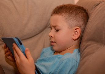  White child plays in the tablet. A boy in a blue T-shirt is sitting on the couch.