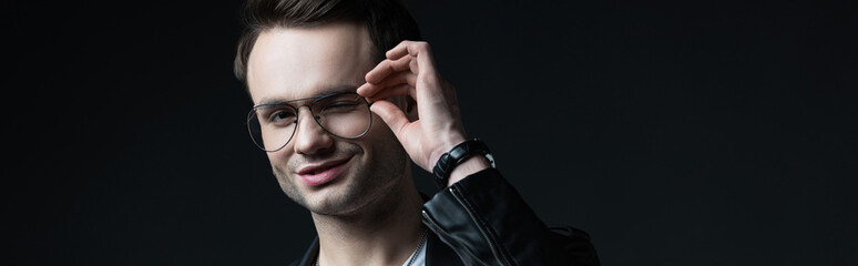 smiling stylish brutal man in biker jacket touching eyeglasses isolated on black, panoramic shot