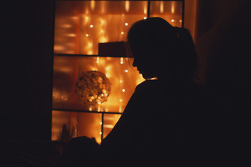 silhouette profile young woman figure sitting in a room decorated with a light garland against shelves with gift boxes and decor , concept evening relaxation, mood and solitude