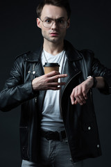 stressed stylish brutal man in biker jacket with wristwatch and coffee to go isolated on black