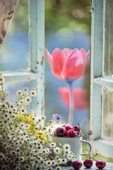 Cherry in an old iron mug and a bouquet of wildflowers on an old wooden window. Vintage photo. Outside, lushly blooming flowers in the garden. Summer mood is a magical photo. summer in the village of 