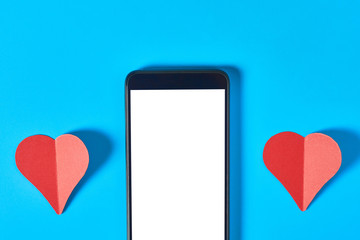 Black smartphone with isolated white screen for text, picture, photo and other graphics near two red paper hearts lies on blue table. Valentines day and love concept. Space for text. Top view