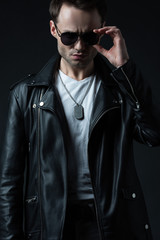 stylish brutal man posing in biker jacket and sunglasses isolated on black