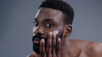 Close-up of handsome young sexy shirtless man applying moisturizing cream on face looking at camera. Nude portrait of attractive guy caring for his skin preparing for a day. - obrazy, fototapety, plakaty